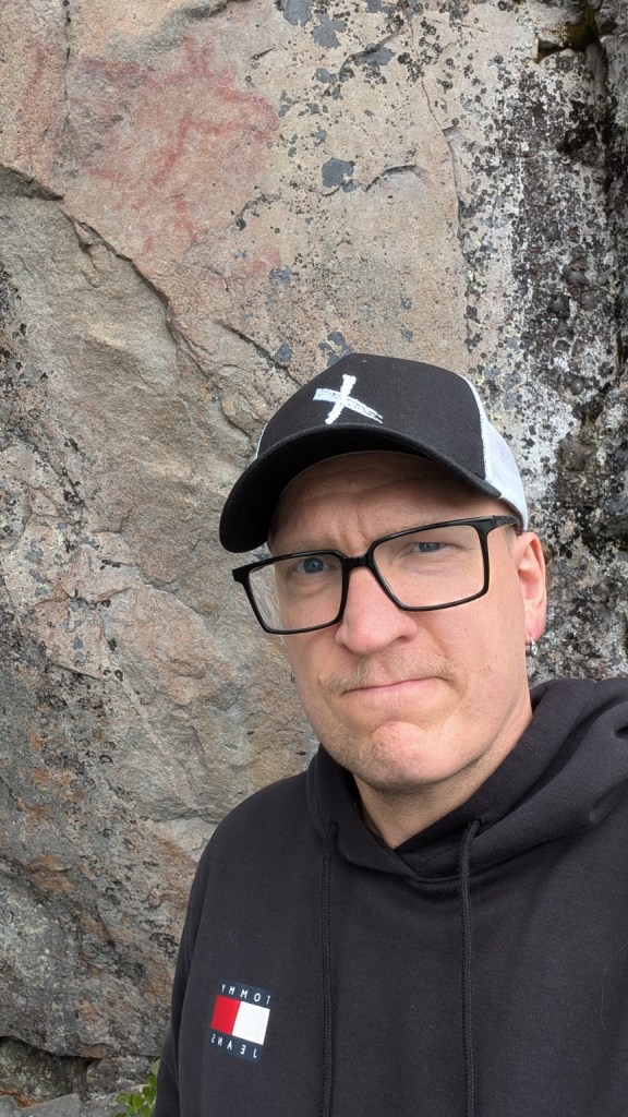 Simo Marttinen silmälaseissa, mustassa lippalakissa ja hupparissa kallion edessä.